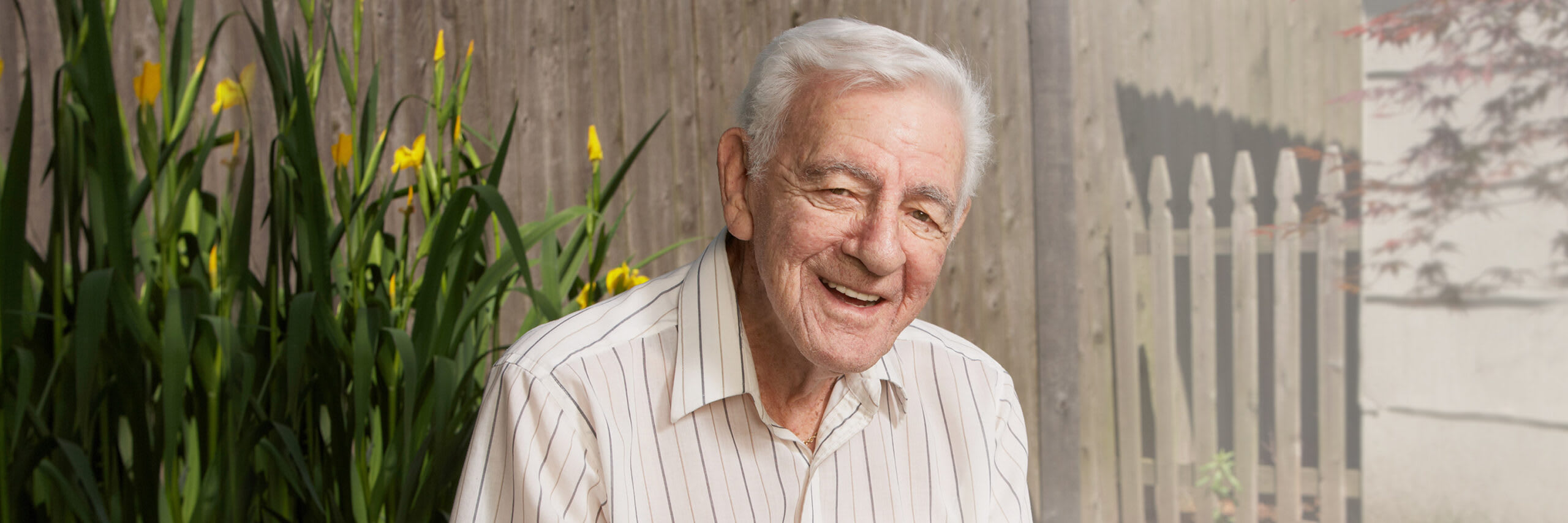 A senior man smiling in front of flowers outside his house