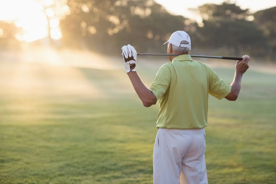 A senior man stands with a golf club draped over his shoulders.