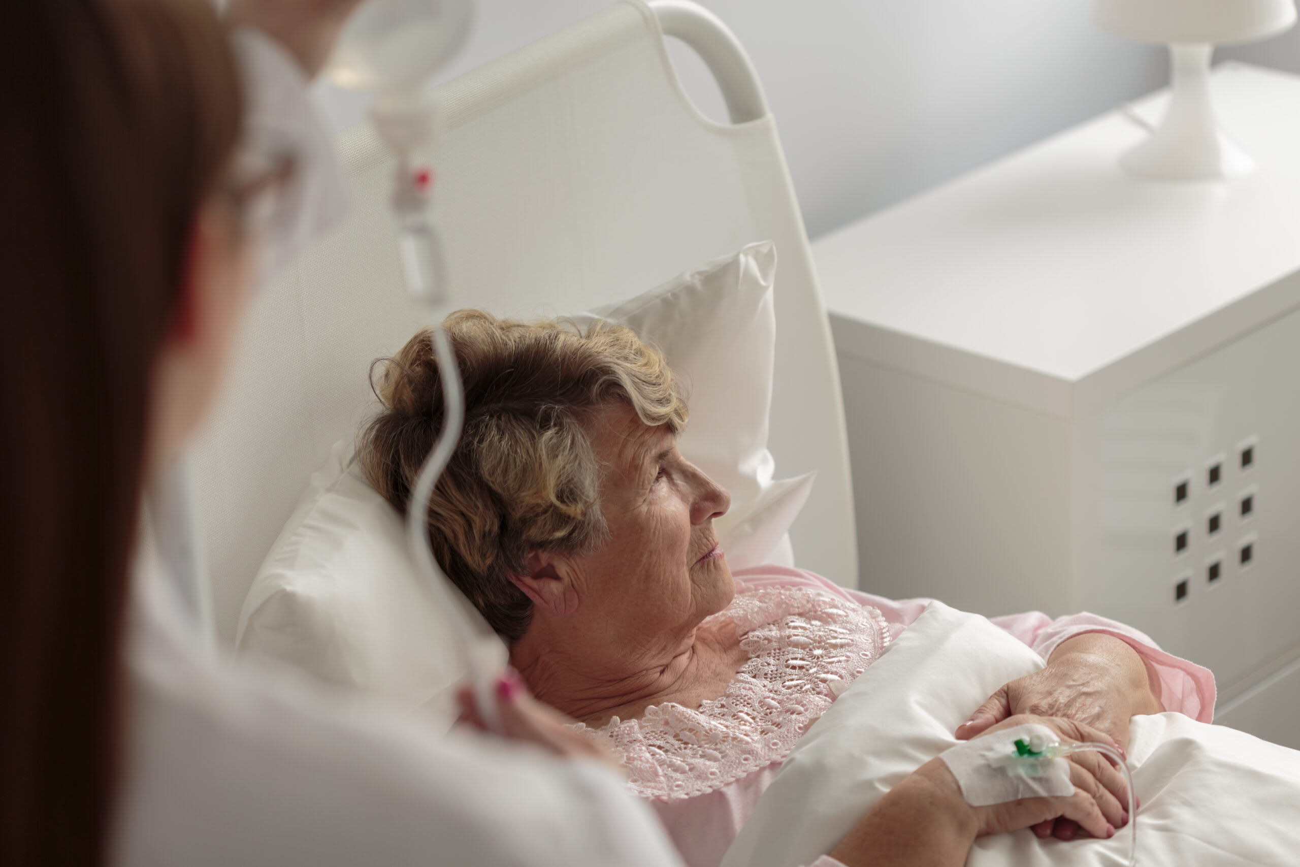 Senior woman sits in hospitable bed with nurse nearby.